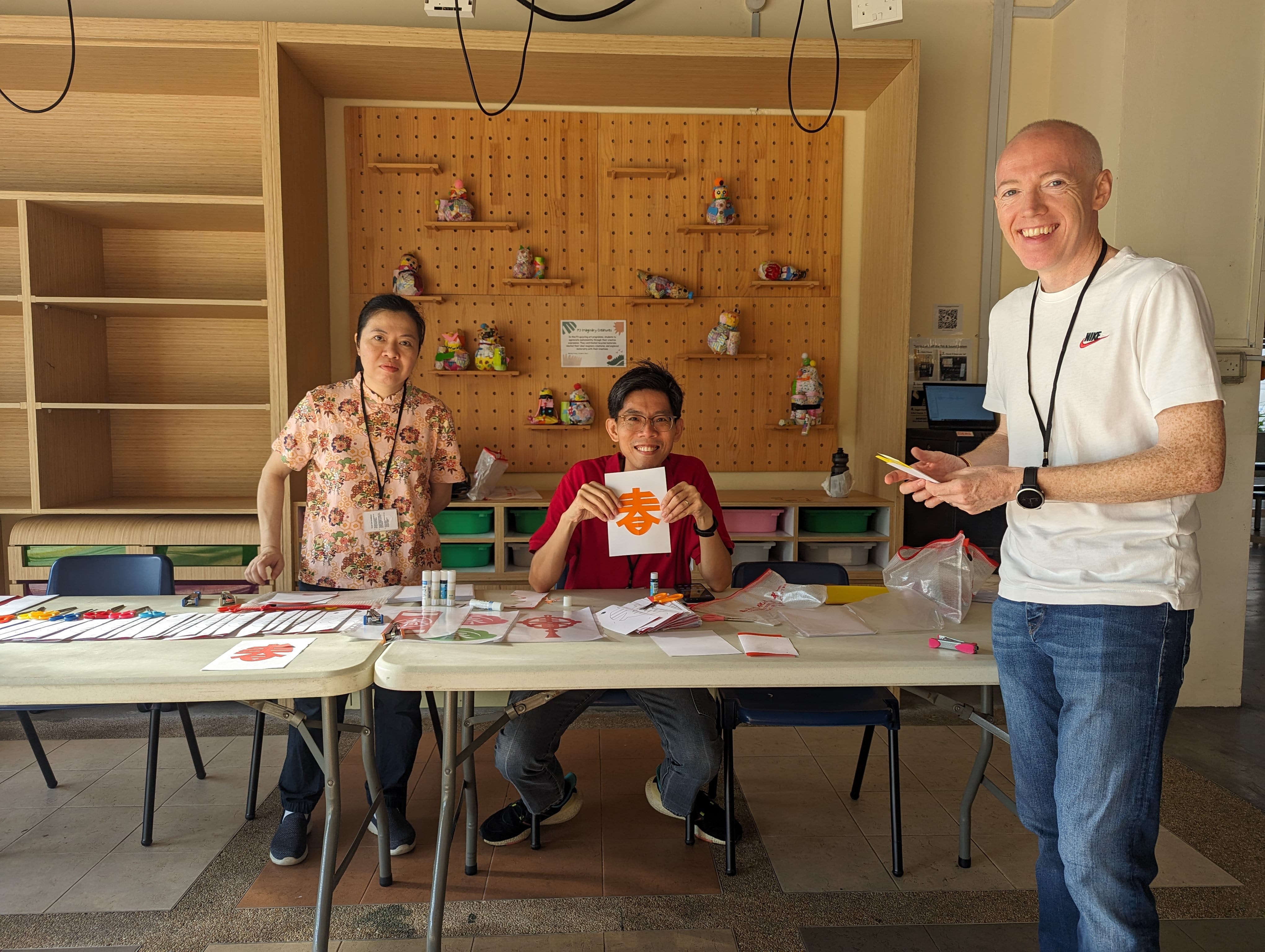 Three people at craft tables; center holds orange Chinese character paper while man on right smiles.